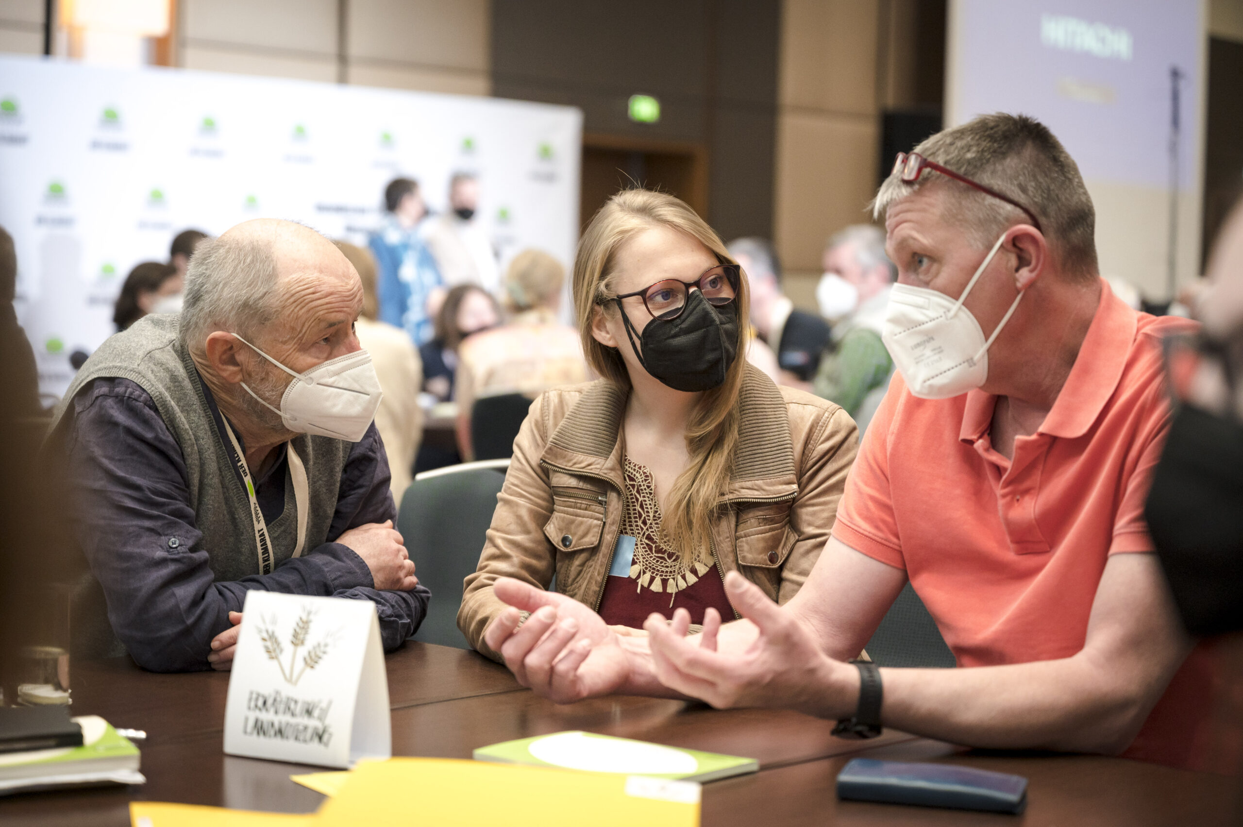 3 Bürger:innen mit FFP2 Masken in Diskussion am Tisch 